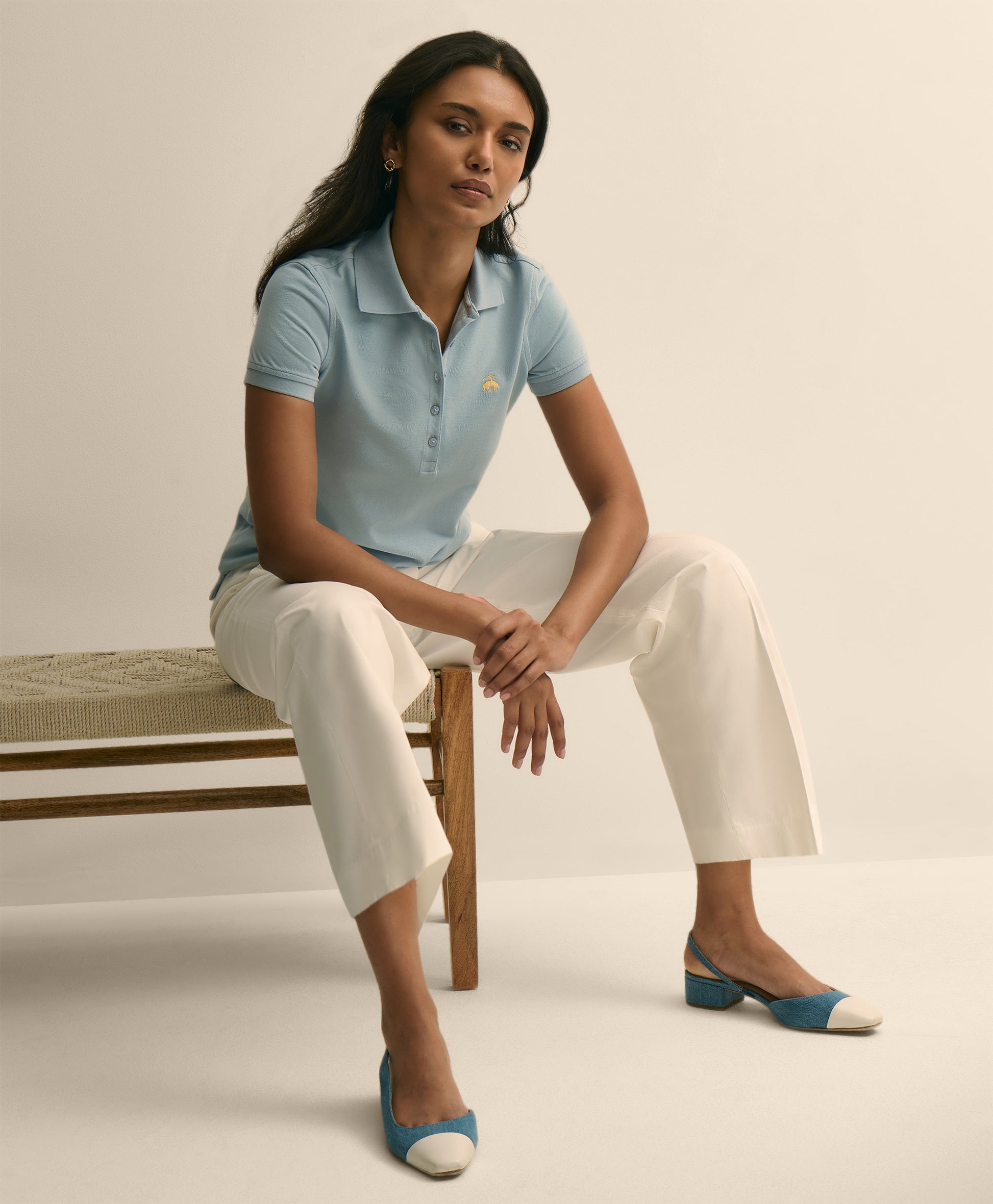 Woman sitting on a bench wearing a light blue polo shirt and white pants.