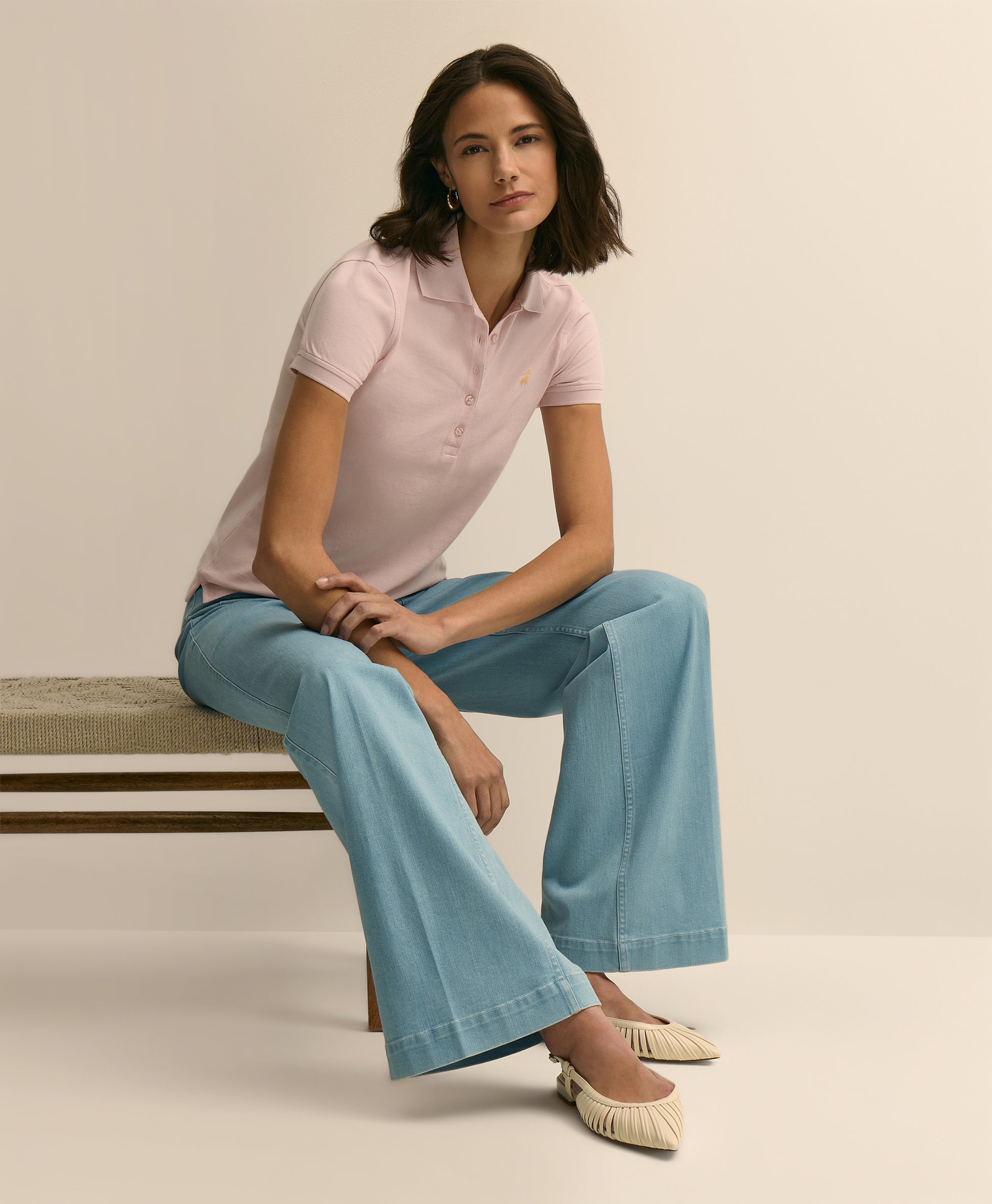 Woman wearing a pink polo shirt and light blue jeans sitting on a bench.