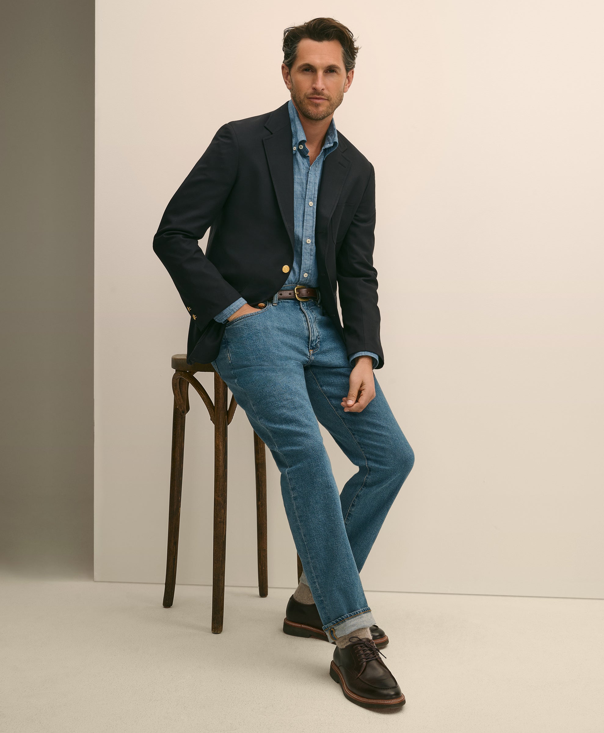 Man wearing a dark blazer over a denim shirt, paired with jeans and brown shoes, leaning against a stool.